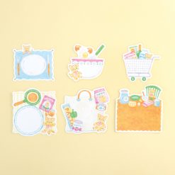 Individual Papier Platz sticker designs laid out on a yellow background, showing teddy bear illustrations with plates, mixing bowl, shopping trolley, grocery bag, food packaging, and kitchen items in a cosy illustrated style.