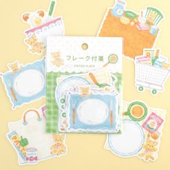 Papier Platz sticker flakes pack featuring a teddy bear in a kitchen and grocery theme, with plates, cooking tools, shopping bag, groceries and pantry items, arranged around the packaging on a soft yellow background.
