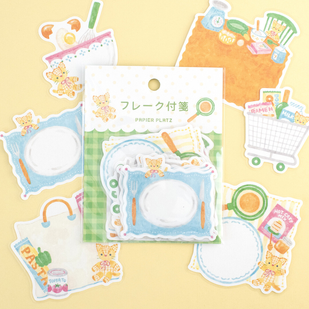 Papier Platz sticker flakes pack featuring a teddy bear in a kitchen and grocery theme, with plates, cooking tools, shopping bag, groceries and pantry items, arranged around the packaging on a soft yellow background.