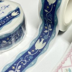 Papier Platz die-cut washi tape unrolled on a white background, showing a blue ribbon-style lattice pattern with small floral details and scalloped edges, alongside the rolled tape.