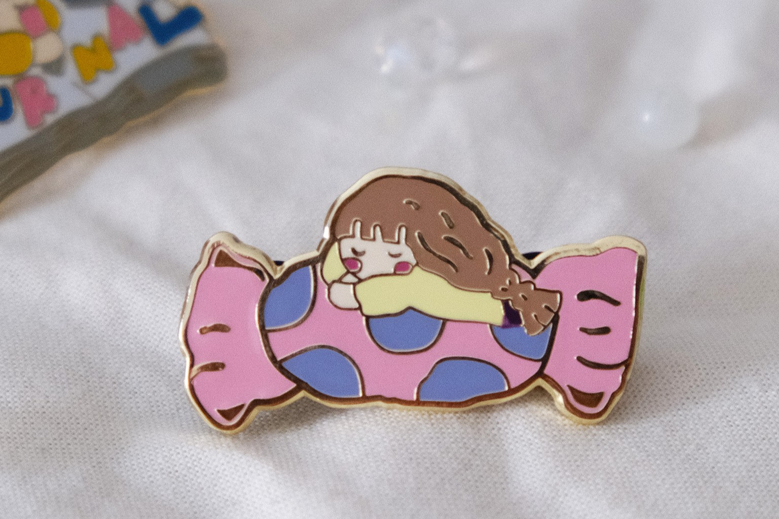 Close-up styled photo of an Eileen Tai sweet like candy enamel pin on a soft fabric background.