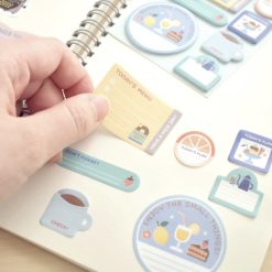 Close-up of a hand holding a mini stationery sticky note from the Eric Small Things set, with additional sticky notes visible in the background on a wood tabletop.